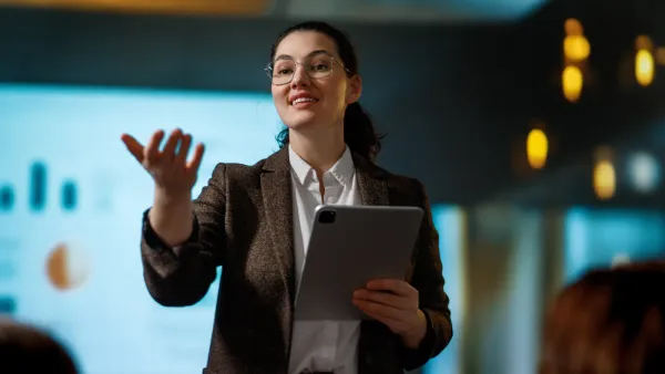 Professional woman confidently presenting with a tablet in a boardroom, leading an executive meeting with data charts on screen behind her