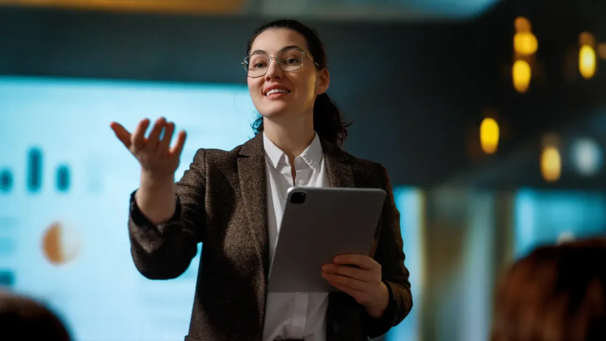 Professional woman confidently presenting with a tablet in a boardroom, leading an executive meeting with data charts on screen behind her